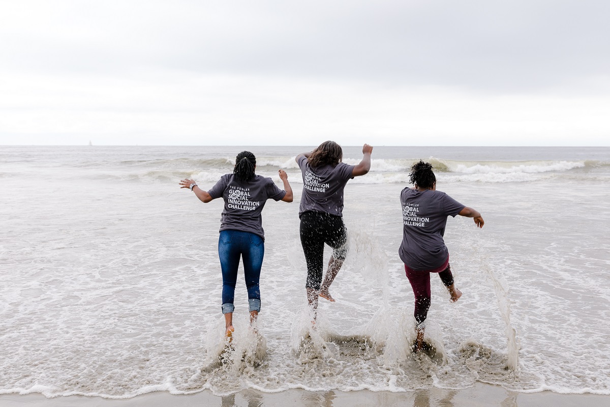 Three women wearing Fowler Global Social Innovation Challenge t-shirts splash in the ocean.
