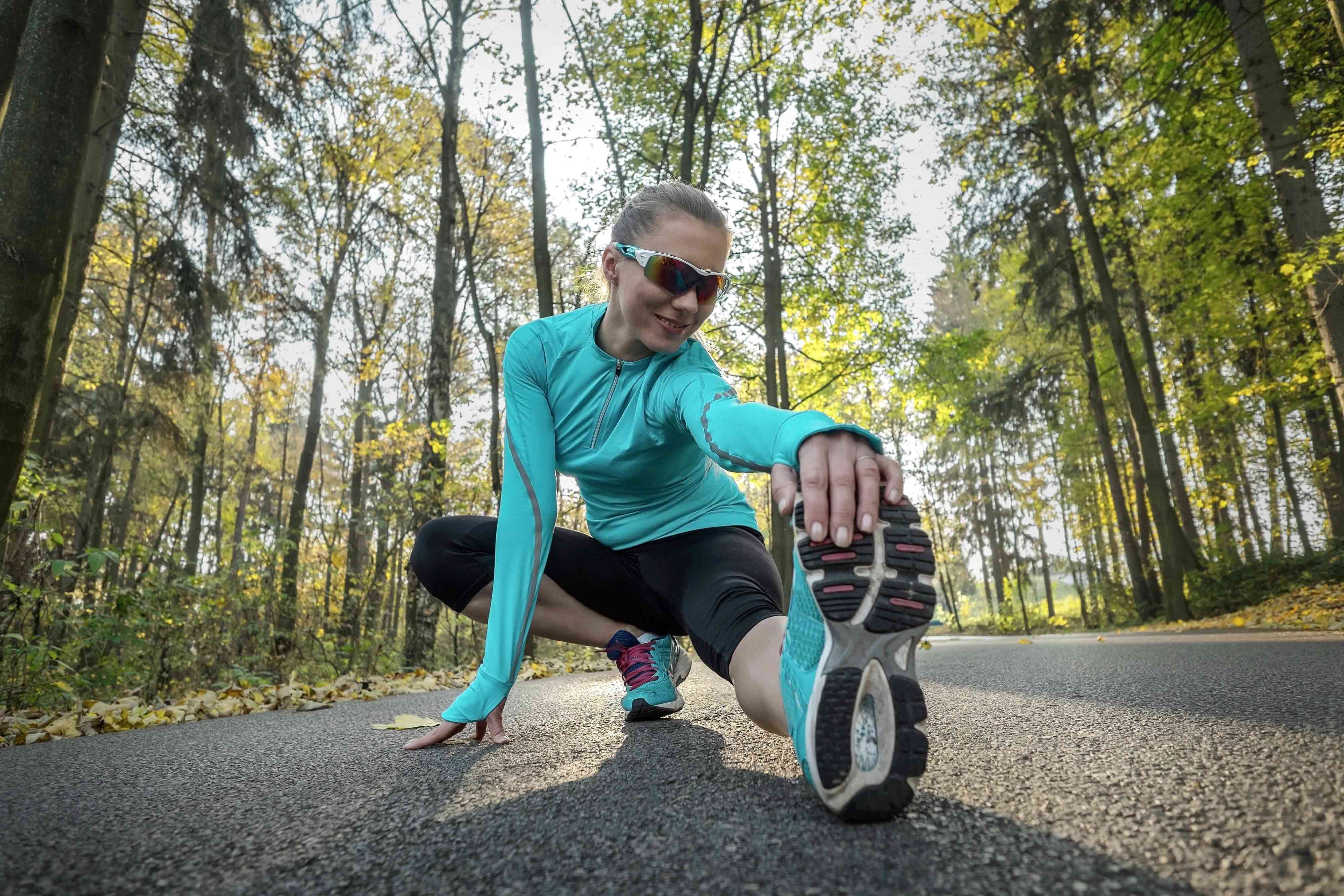 A person in running clothes on a tree lined path, stretching her leg. 