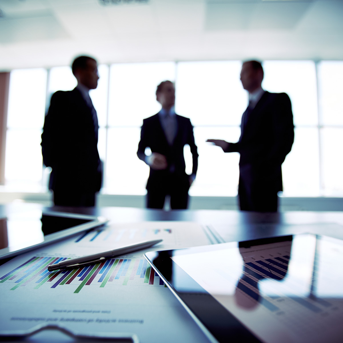 Graphs and Tablets on a table with three people in business suits speaking in the background.