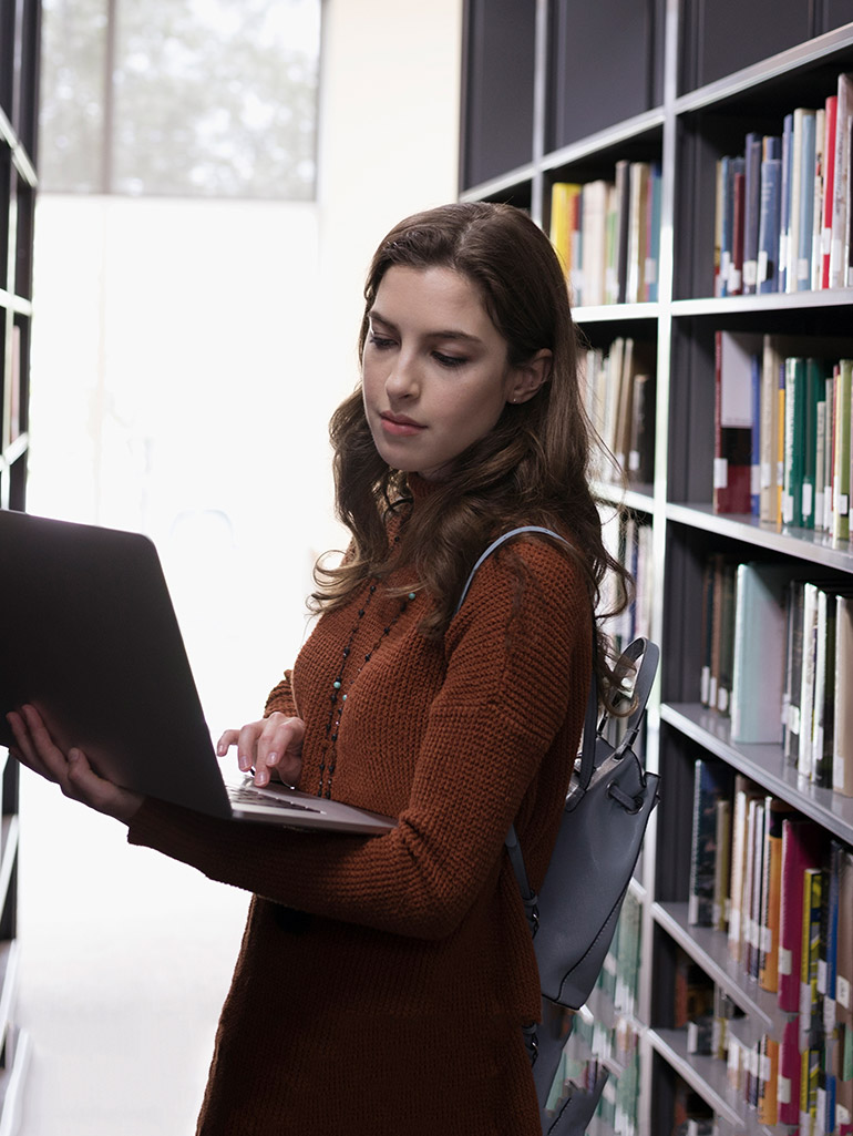 A student standing between bookshelves using a laptop in the library.