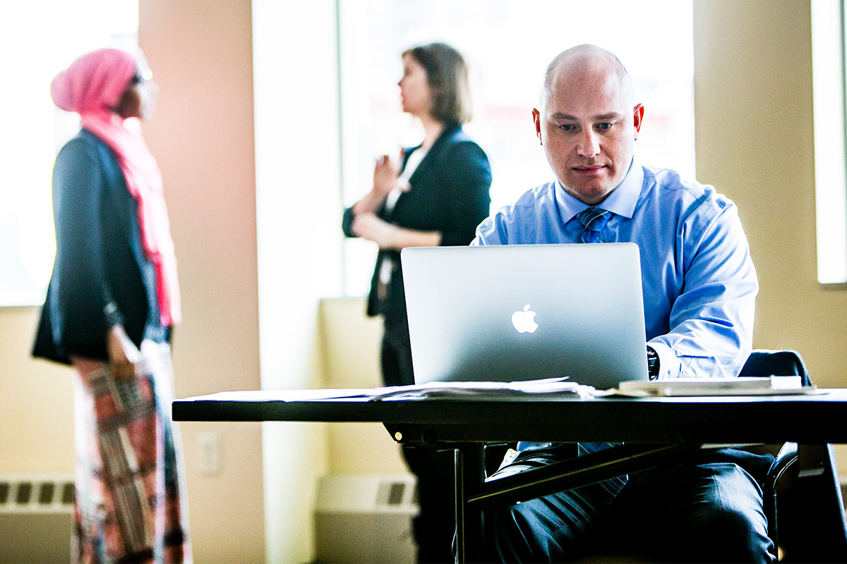 A student sits with a laptop computer as two other students have a discussion in the background.