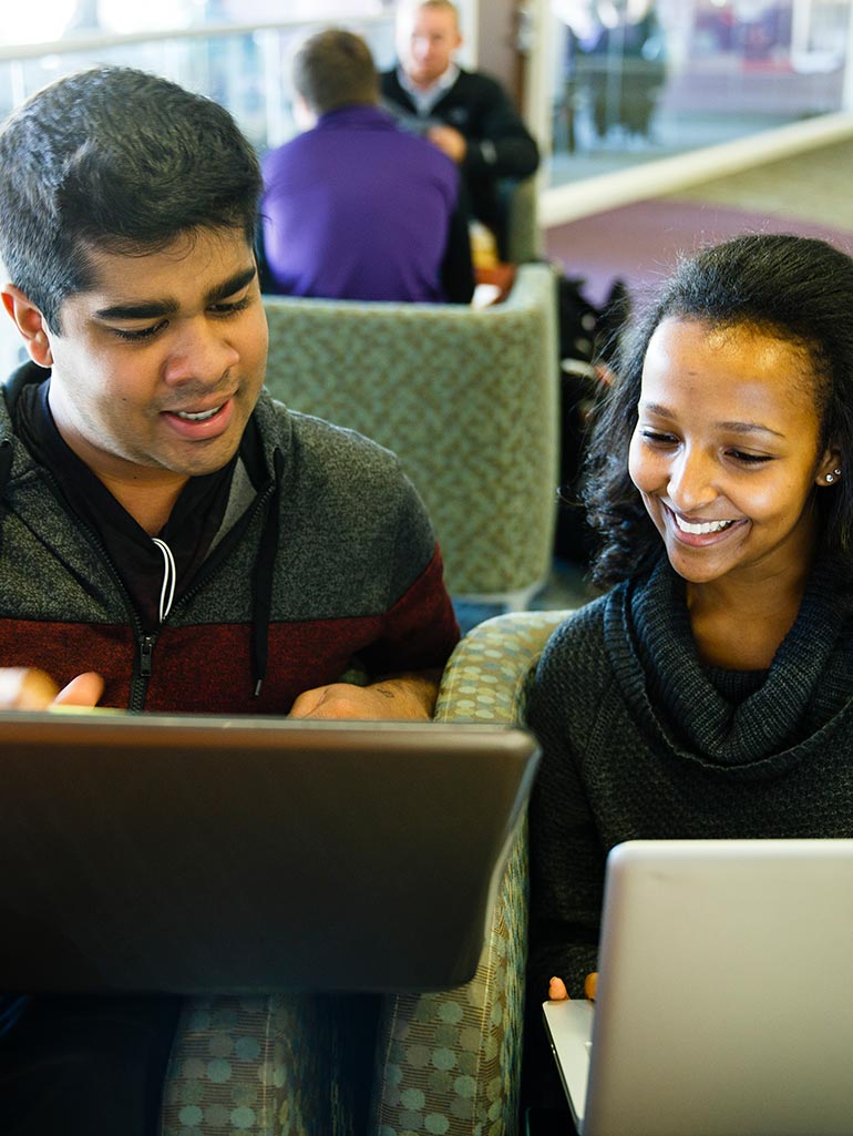 Two students smile as they work on their laptops while sitting on a couch.